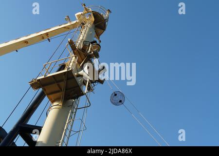 View on forward vessel mast with one black anchor ball. There are also jib of ship's crane, ladder and platform with foghorn. Behind is clear blue sky Stock Photo