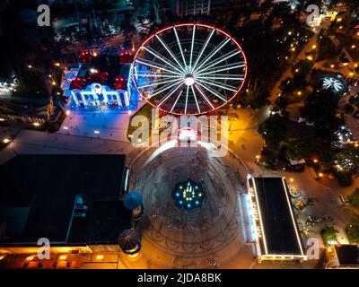 Aerial look down on illuminated Ferris wheel with fountain and attractions. Kharkiv city center recreation area in evening lights. Amusement Gorky Cen Stock Photo