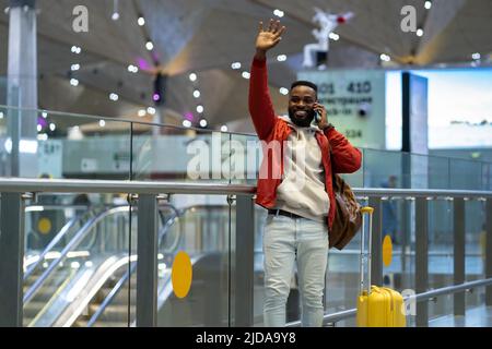 Young joyful african man traveler waiting friends in airport, waving hand and talking by phone Stock Photo