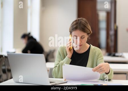 Middle-aged woman busy with documents talking on cellphone working in campus open space. Education.  Stock Photo