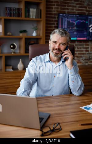 Portrait of smiling businessman talking on the phone using laptop in office interior woth stock market graphs on background. Male trader negotiates Stock Photo