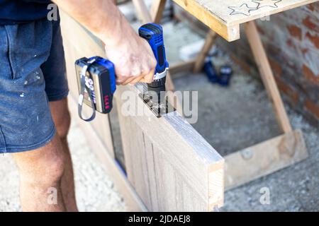 A carpenter is using an electric drill to screw the hinge hardware onto a wooden door. Door installation. Stock Photo