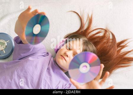 Happy teenage girl in lilac t-shirt lies on bed in pink headphones holds cd disks in her hands Stock Photo