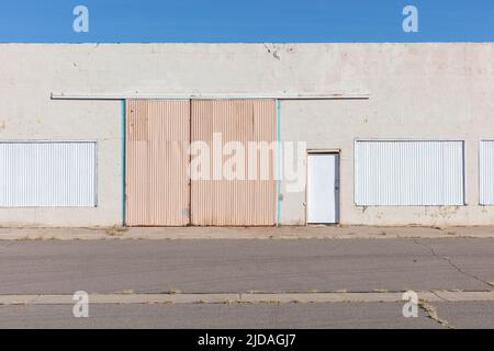 Closed warehouse building with metalwork shutters on doors and windows, and weeds growing through tarmac. Stock Photo