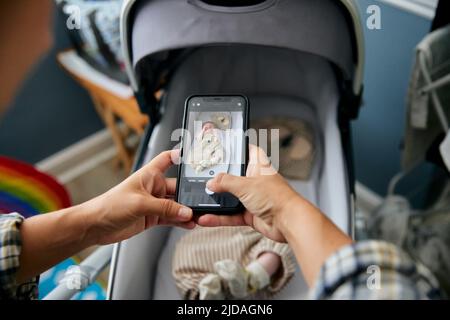 Mother taking photograph of 3 month old baby boy lying in baby buggy using smart phone Stock Photo