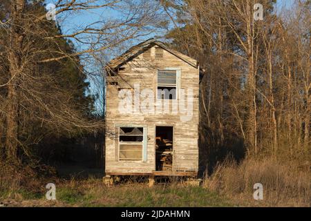 A rural homestead or small house abandoned and crumbling, overgrown with plants and shrubs.