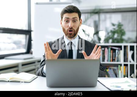 Excited successful caucasian man, businessman, manager, economist, sitting at a work desk in front of a laptop screen, having a video consultation with colleague, pleasantly surprised by the good news Stock Photo