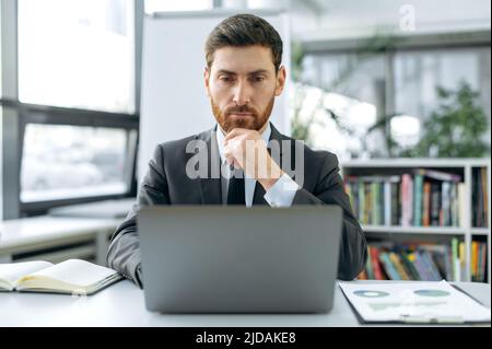 Focused intelligent Caucasian male, marketer, web developer, financial manager, sits at a desktop in front of a laptop, in a modern office in stylish business suit, concentrated working in a laptop Stock Photo