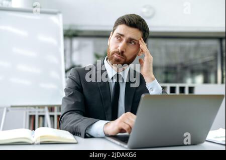 Pensive Caucasian male with a beard, director of company, top manager, sits at a desk in front of a laptop in modern office, looks thoughtfully away, analyzes a work task, thinks over a strategy Stock Photo