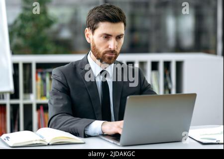 Focused handsome smart caucasian businessman, lawyer, auditor, IT specialist, sits in modern office, using laptop, working on a new project, texting with client, answer email,analyzes the stock market Stock Photo