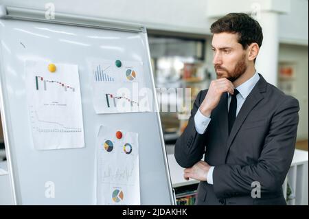 Focused elegant young adult caucasian man, company top manager, financial analyst, risk manager, stands near whiteboard with graphs in a modern office, concentrated looks at them, analyzes dynamics Stock Photo