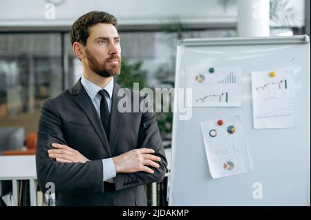 Portrait of elegant successful caucasian attractive man in a business suit, business leader, standing near marker board with financial charts, in a modern office, looking thoughtfully to the side Stock Photo