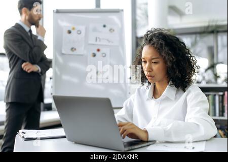 Focused African American woman, sales manager, marketer, sitting at a desk in front of a laptop. In the background, a Caucasian colleague, stands near a whiteboard with graphs, analyzes the dynamics Stock Photo