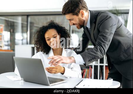 Positive smart multi ethnic colleagues, caucasian man and african american girl, web developers, in modern office working on laptop, writing an algorithm for a mobile application, discussing details Stock Photo