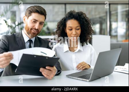 Positive successful work colleagues, a caucasian man and an african american woman, are sitting in office, working on a laptop and with documents, discussing and analyzing data, compiling reports Stock Photo