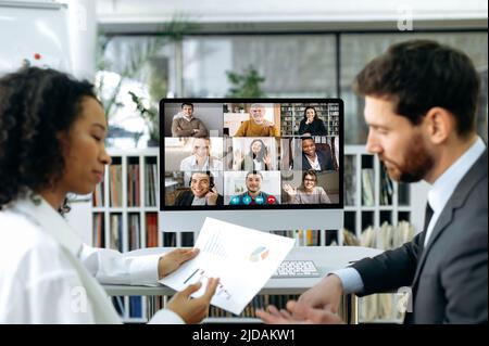 Company co-founders, a caucasian man and an african american woman, sit in a modern office in front of a computer screen, have an online video meeting with colleagues, demonstrate company profit Stock Photo