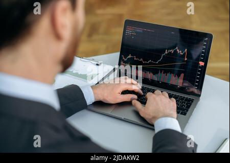 Trading the stock market. Successful smart caucasian businessman, stock market trader, sits at his desk, uses a laptop, works with quote charts, assesses risks and possible profits,develops a strategy Stock Photo