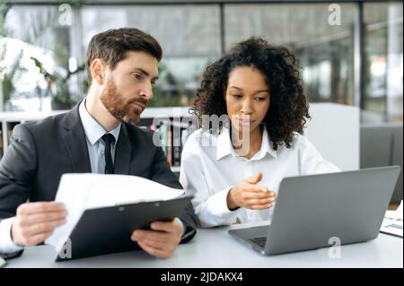 Business colleagues, an African American woman and a Caucasian man, company owners sitting at a desk in office, working on a laptop and with financial documents, preparing a contract for cooperation Stock Photo