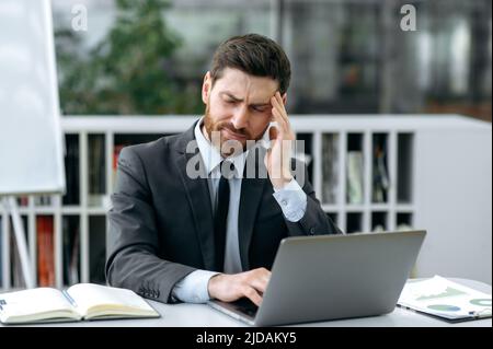 Exhausted overworked sad caucasian businessman, company owner, financial advisor, manager, sitting at a desk in front of a laptop, having migraine from online work, holding his head, needing rest Stock Photo