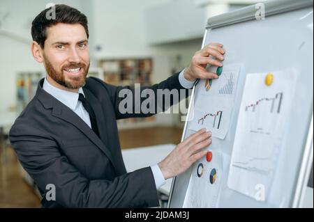 Smart successful handsome caucasian male, financial business leader, pinning charts on a whiteboard, while standing in a modern office, in a formal suit, looking at the camera and smiling friendly Stock Photo