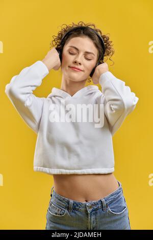 Smiling calm girl in modern headphones with closed eyes enjoying song. Portrait view of pretty woman with black earphones listening music, having rest, isolated on orange background. Concept of relax. Stock Photo
