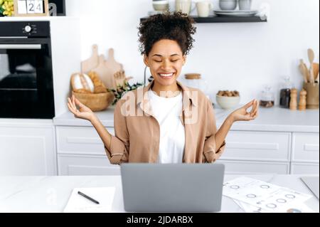 Satisfied peaceful African American curly haired girl, freelancer or student, taking a break, resting from online work or study in a laptop, meditating with closed eyes, relaxing, smiling Stock Photo