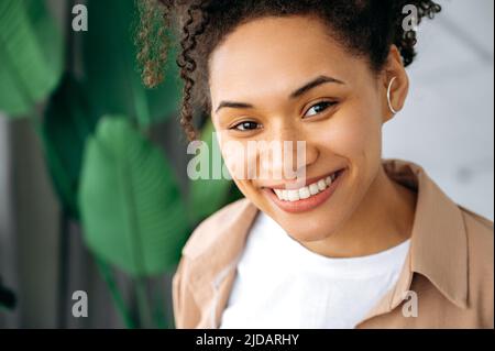 Close-up photo of a positive gorgeous African American curly haired girl, mixed race female with freckles and perfect white manicured teeth, looks directly at the camera, with happy toothy smile Stock Photo