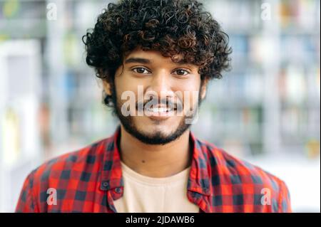 Close-up photo of curly-haired confident positive mixed race guy, Indian or Arabian male, in casual wear, manager, designer, student or freelancer, stands indoors, looks at camera, smiling friendly Stock Photo