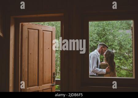 View from inside the cottage. Romantic groom kissing brunette bride on the balcony. Romantic moment. Wedding Day concept Stock Photo