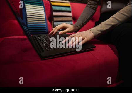 Details: Hands of unrecognizable woman typing text on laptop keyboard, sitting on a red velour sofa with colorful upholstery fabric samples on backgro Stock Photo