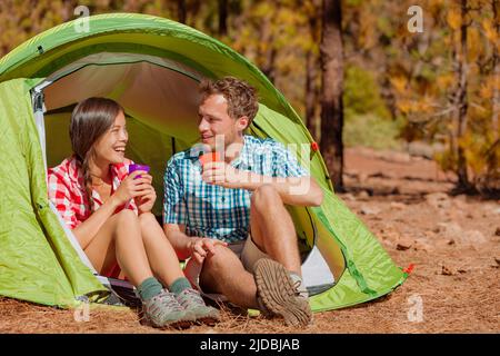 Camping couple drinking water in tent smiling happy outdoors in forest enjoying sun at looking at view. Happy multiracial couple relaxing after Stock Photo
