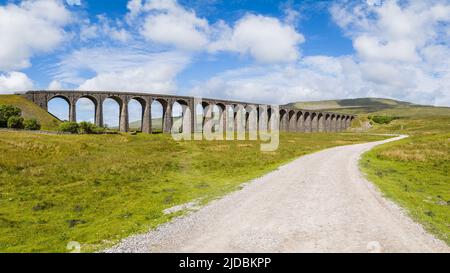 Multi image panorama of a pathway leading to the Ribblehead Viaduct in North Yorkshire seen under a bright blue sky.