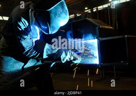 A welder in protective overalls and a welding mask welds metal structures by welding on a semi-automatic line, blue sparks fly to the sides Stock Photo