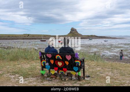 Two people on a bench that has been the subject of 'craft bombing', with a view of Lindisfarne Castle from harbour on Holy Island, Northumberland, UK. Stock Photo