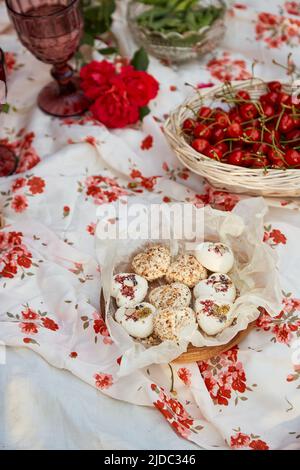 Aesthetic summer picnic in the sunny backyard. Tablecloth, box of marshmallows, strawberries, cherry, peas and glasses of wine among pink roses. Romantic evening, appreciation of the moment Stock Photo