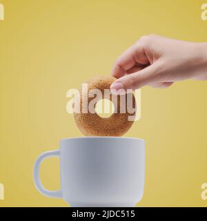Woman dipping a donut shaped cookie in a cup, she is having breakfast or a snack, copy space, isolated on yellow background Stock Photo