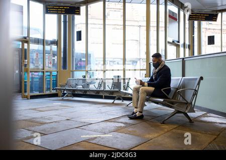 Man wearing face mask sitting on bench in bus terminal using mobile Stock Photo