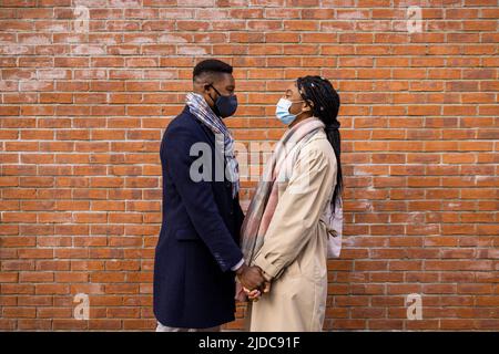 Couple with face coverings standing face to face by brick wall and holding hands Stock Photo