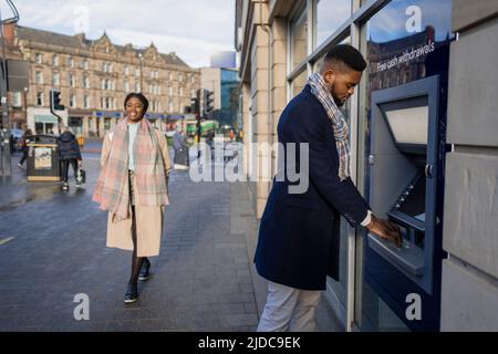 Man withdrawing money from atm as woman walks past in city centre Stock Photo