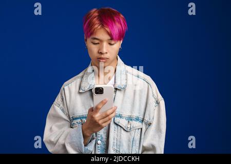 Asian boy in denim jacket posing with mobile phone isolated over blue background Stock Photo