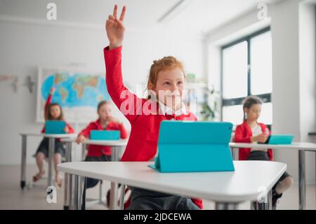 Happy schoolgirl with hands up using digital tablet during lesson in classroom at primary school. Stock Photo