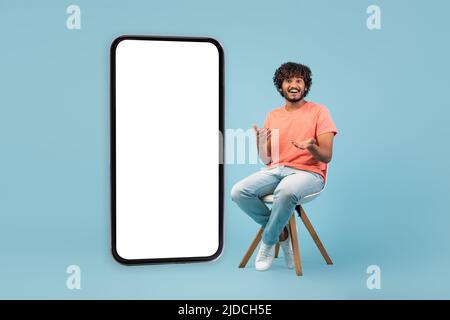 Amazed hindu guy sitting on chair by huge smartphone, mockup Stock Photo