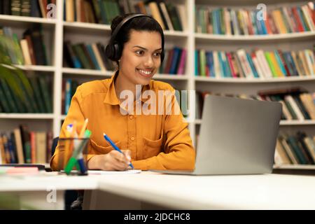 Modern technology and elearning concept. Cheerful lady in headset watching webinar online, sitting in library Stock Photo
