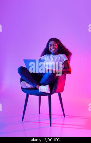 Full length of charming young black woman working online, sitting in armchair and using laptop in neon light Stock Photo