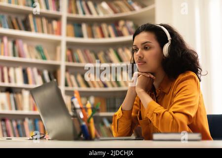 Pensive student lady looking at laptop computer, learning online, having remote class with distant teacher, sitting in library Stock Photo