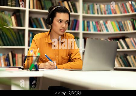 Young mixed race lady having online class, sitting at desk in library in front of laptop, using headset and taking notes Stock Photo