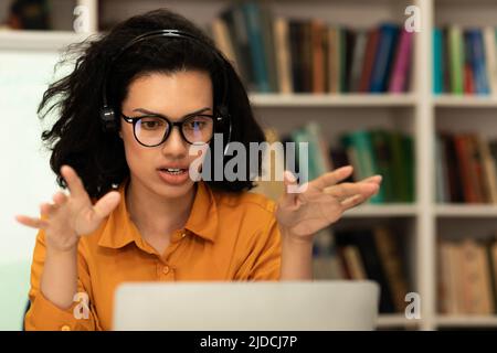 Modern remote education. Female teacher having videochat with students, talking to laptop webcam, sitting in library Stock Photo