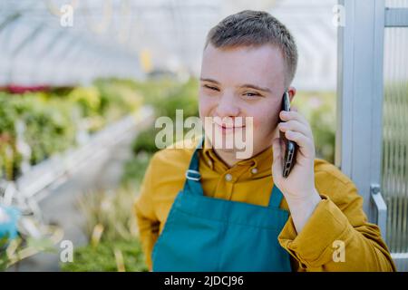 Young employee with Down syndrome working in garden centre, standing in door of greenhouse and calling on cellphone. Stock Photo