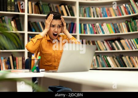 Shocked mixed race woman sitting in library, staring at laptop screen, studying or working remotely Stock Photo