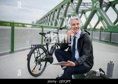 Businessman with bike sitting on bridge, using tablet and listening to music. Commuting and alternative transport concept Stock Photo